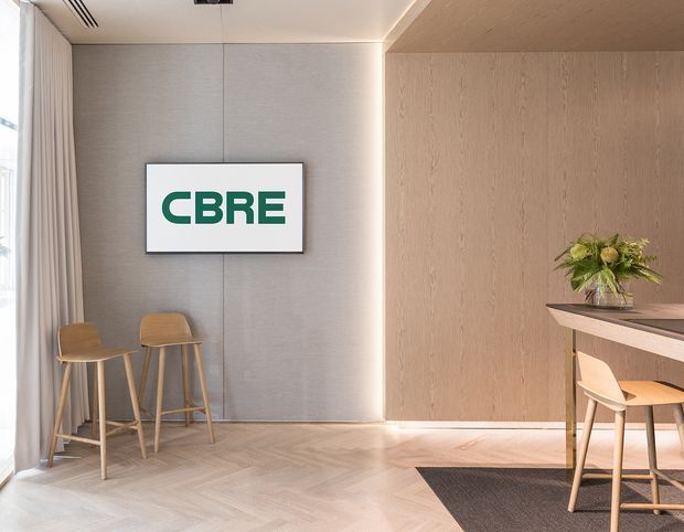 A refined commercial reception or waiting area with a CBRE sign mounted on a textured grey wall. The space features pale timber wall panelling, herringbone timber flooring, a slim reception counter with timber finish, bar stools, soft sheer curtains, integrated linear lighting, and a small floral arrangement. The palette is warm and understated with contemporary detailing.