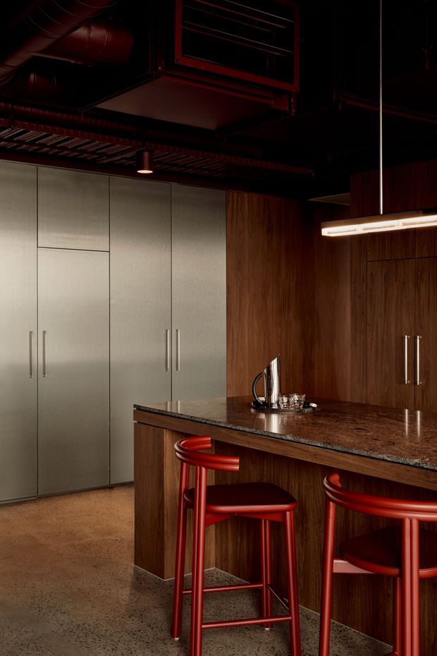 A dramatic, dark-toned kitchen and bar area with a large central island, integrated tall cabinetry, richly grained dark timber joinery, and a mottled stone or terrazzo benchtop. Deep red bar stools and linear pendant lighting provide bold accents. The exposed dark ceiling services and ductwork give the room an industrial character, while warm low lighting creates a moody, sophisticated atmosphere.