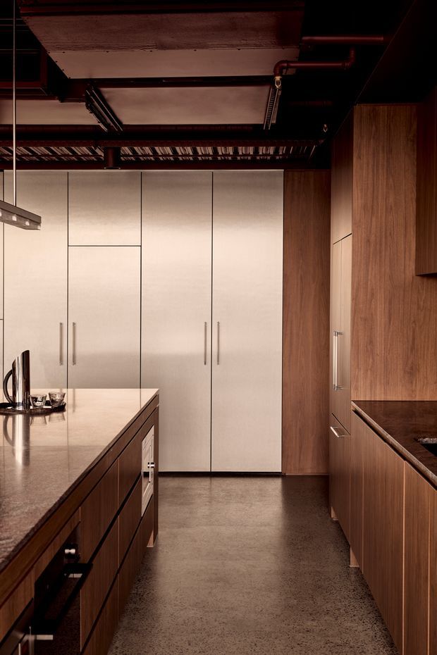 A sophisticated galley-style kitchen with a large central island, tall pale integrated cabinetry, and warm walnut-toned cabinetry along the side wall. The kitchen has dark exposed ceiling services and beams, slender linear lighting, stone benchtops, and a polished concrete or terrazzo-like floor. The restrained palette and dramatic low lighting create an upscale industrial-modern atmosphere.