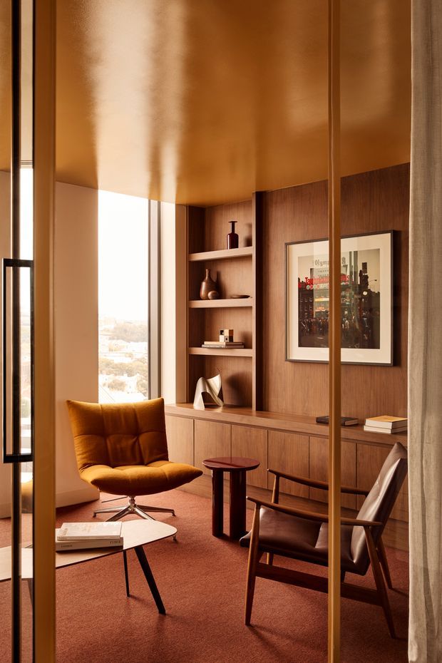 Warm, refined interior vignette viewed through slim bronze-toned glazed framing and curtains. The room features a built-in dark timber shelving and cabinetry wall, floor-to-ceiling glazing, a rust-orange upholstered lounge chair, leather armchairs, small timber side tables, and a pale stone or light tabletop coffee table. The ceiling and walls have rich caramel and terracotta tones, creating an intimate, luxurious mid-century-inspired atmosphere.