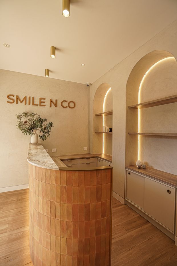 Warm, softly lit reception area for “SMILE N CO,” featuring a curved terracotta-tiled reception desk with a pale stone benchtop, timber flooring, textured beige walls, and built-in arched shelving with integrated LED lighting. Brass-toned pendant and recessed lighting create a refined, welcoming atmosphere.