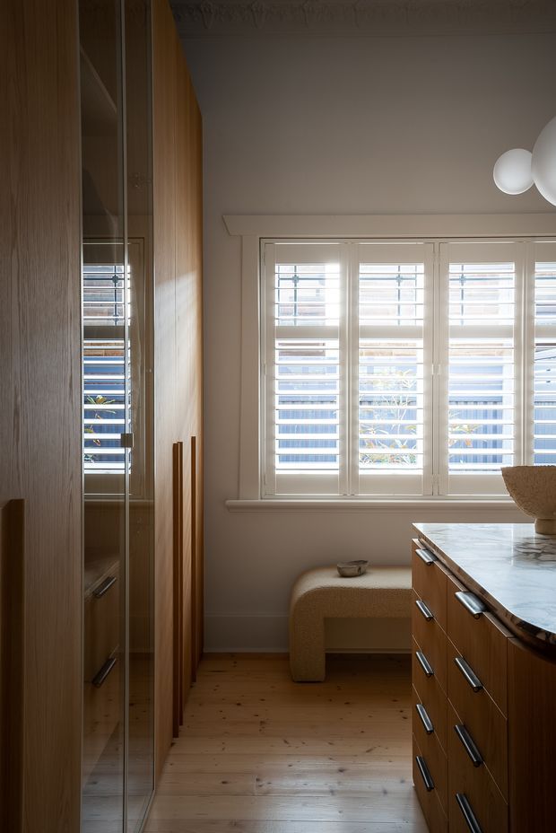 A warm, compact kitchen interior featuring full-height timber cabinetry with integrated handles, a reflective glass-fronted cabinet, a pale stone benchtop, and a built-in upholstered window seat. White plantation shutters span the large window, while timber flooring and soft neutral finishes create a refined, intimate atmosphere.