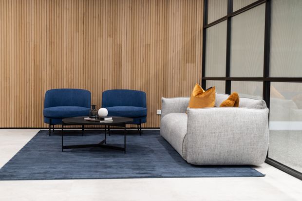 A refined contemporary lounge with two deep-blue upholstered armchairs and a large light-grey sofa arranged around a slim black coffee table. The room features a full-height vertical timber-slat feature wall, expansive black-framed glazing, a pale floor, and a dark blue area rug, creating a calm, modern meeting or living space.