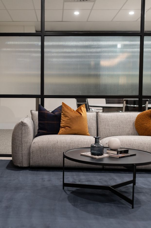 Contemporary lounge area featuring a large light-grey upholstered sofa with navy and ochre cushions, a black round coffee table, and dark flooring. A black-framed internal glazed partition with ribbed translucent glass separates the seating area from an office or meeting space beyond. The scene has a refined, modern commercial-interior feel with muted tones and warm accent cushions.