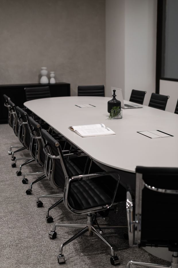 A modern conference room with a large pale oval meeting table surrounded by black leather office chairs. The room has grey carpet, dark built-in cabinetry, neutral textured walls, a large window, and minimal tabletop accessories including documents and a small plant.