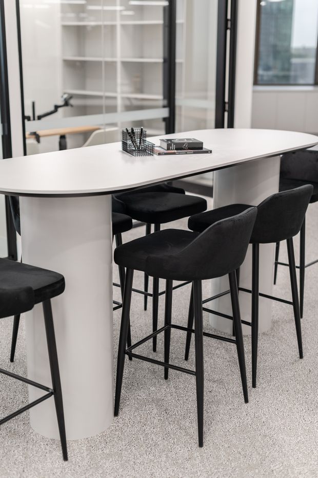 A contemporary office or meeting area with a large white oval communal table supported by cylindrical pedestal legs, surrounded by black upholstered bar-height stools. Black-framed glass partitions reveal shelving and a workspace beyond. The room has a light speckled carpet and a clean monochromatic palette.