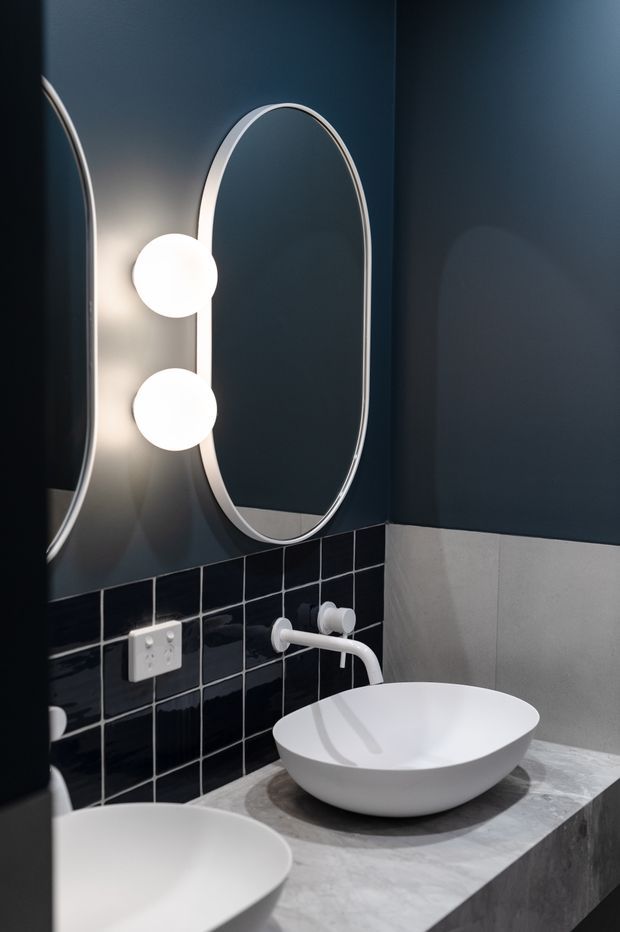 A moody contemporary bathroom vanity featuring a grey stone benchtop, white vessel basins, a wall-mounted white tap, glossy black square wall tiles, and deep navy painted walls. An oval mirror with integrated spherical wall lights adds a sculptural focal point.