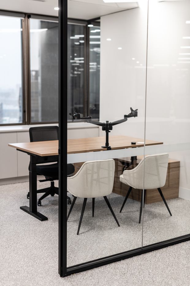 A compact contemporary office meeting room enclosed by black-framed glass partitions. The room contains a timber meeting table with a central monitor arm, two cream upholstered visitor chairs, and a black office chair. White built-in cabinetry, large windows, recessed ceiling lighting, and light grey carpet create a bright, clean workspace.
