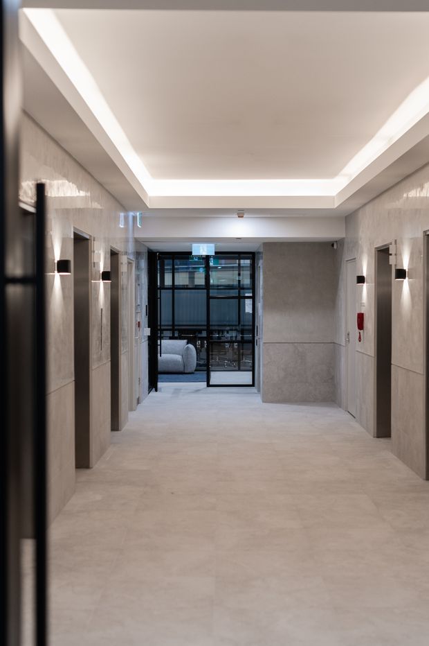 A refined contemporary interior hallway with a wide, symmetrical corridor finished in pale beige-grey stone or concrete-look surfaces. Multiple recessed doorways and dark wall-mounted sconces line both sides, while a large recessed ceiling panel with concealed perimeter lighting creates a softly illuminated cove. Black-framed glazed doors at the far end open toward a furnished room beyond.