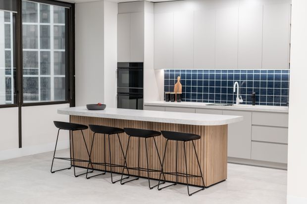 A bright contemporary kitchen in an urban apartment, featuring a large island with a white stone benchtop, vertically fluted timber joinery, four black bar stools, white handleless cabinetry, integrated black appliances, and a glossy dark blue tiled splashback. Full-height black-framed windows overlook a dense city setting.