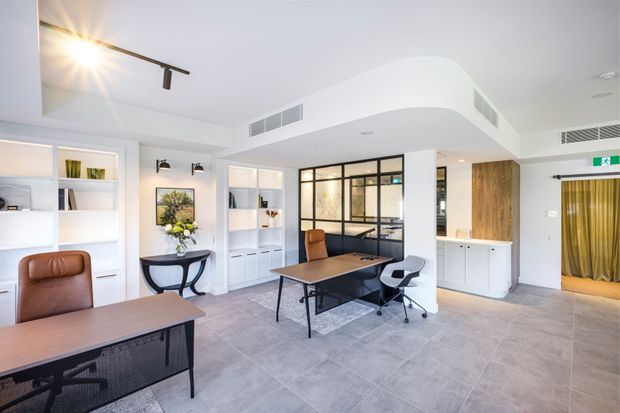 Bright contemporary office interior with several workstations, built-in white shelving and cabinetry, a central desk, glass-and-black-metal framed partition, timber-fronted storage, tiled flooring, and curved white bulkhead ceilings. The space has abundant daylight, recessed and track lighting, and a clean professional fit-out.