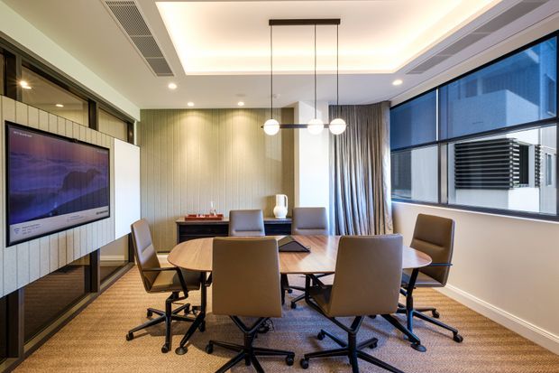 A contemporary meeting room or home office with a large oval timber conference table surrounded by upholstered swivel chairs. The room features a wall-mounted display screen, vertically panelled feature walls, full-height curtains, expansive windows, recessed perimeter ceiling lighting, and three suspended globe pendant lights. The palette is warm and neutral, with beige carpet, light timber tones, taupe upholstery, and dark window and furniture accents.