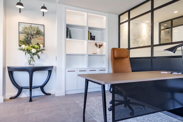 A refined home office with a large wood-look desk, black mesh office chair, tan leather executive chair, black-framed glazed partition, and built-in white shelving. The room has pale walls, large light-grey floor tiles, minimalist pendant lighting, and a console table beneath wall art and floral arrangements.