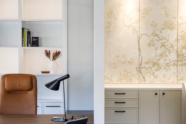 A refined home office or study viewed through a partial partition, with a warm brown leather chair, dark timber desk, black angled desk lamp, white built-in shelving and drawers, and a pale floral-patterned decorative wall panel or wallpaper behind light cabinetry. The space has a restrained, elegant palette and clean built-in joinery.