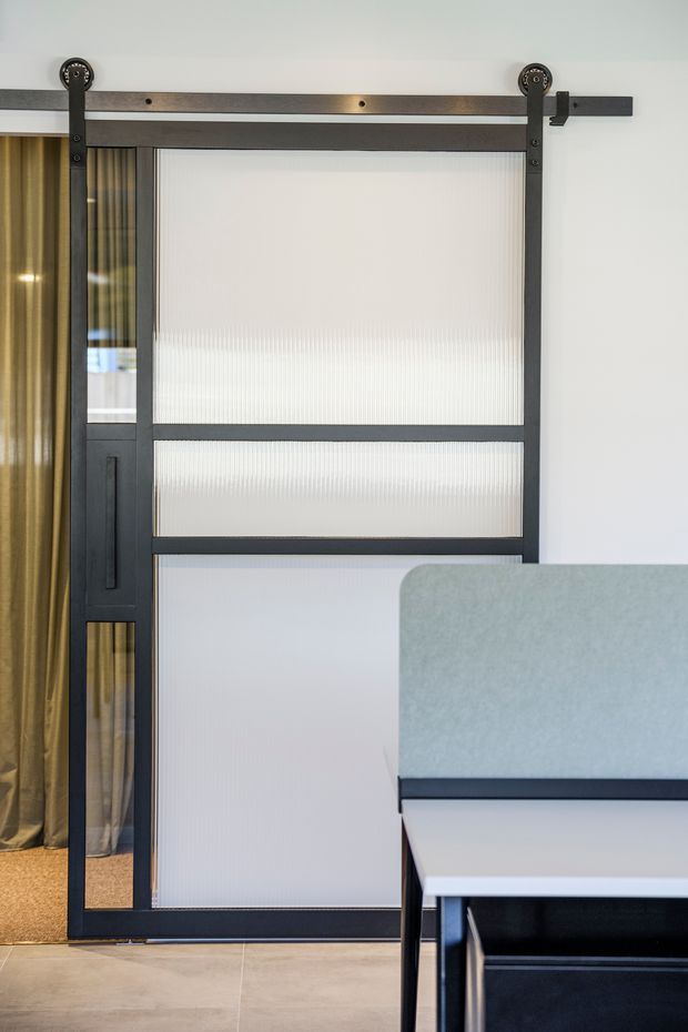 Interior detail featuring a black-framed sliding barn-style door with translucent ribbed glass panels and a dark timber-and-metal overhead track. A warm gold curtain is visible behind the door, with a pale tiled floor and part of a contemporary dining or work table in the foreground.