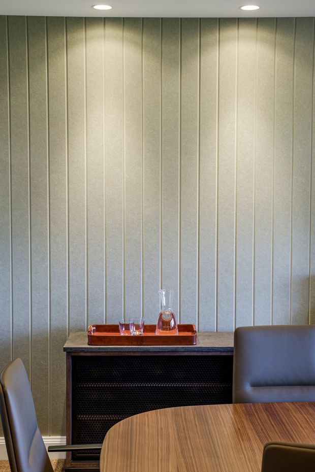 A refined dining or lounge area with a vertically panelled pale grey wall, recessed ceiling downlights, a dark built-in console or fireplace surround, and a round timber dining table with upholstered chairs. A small drinks tray and decanter sit on the console. The space has a restrained, contemporary interior with warm timber and dark contrasting details.