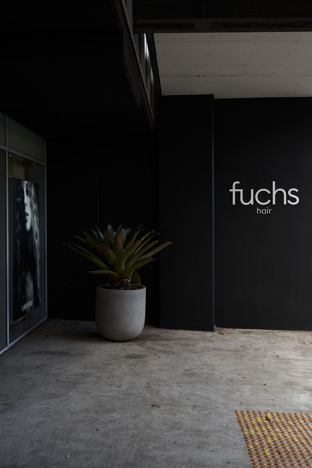 A dark, minimalist commercial hair salon entrance or facade. The black wall features a prominent white “fuchs hair” logo, with dark-framed glazing and a poster at left. A large potted tropical plant sits on a raw concrete floor beneath a light-coloured soffit, and tactile paving is visible at the lower right.