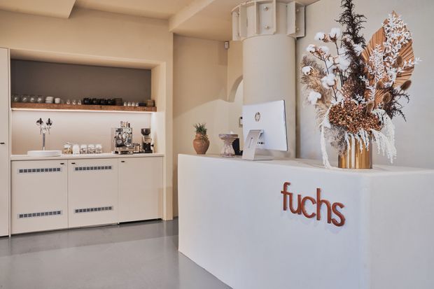 A refined reception and hospitality counter in a softly lit commercial interior. The white curved reception desk features a prominent “fuchs” logo and a large dried floral arrangement in a metallic vase. Behind it is a compact built-in beverage and coffee service area with pale cabinetry, open shelving, cups, and espresso equipment. The space uses warm beige walls, white joinery, smooth grey flooring, and rounded architectural forms.