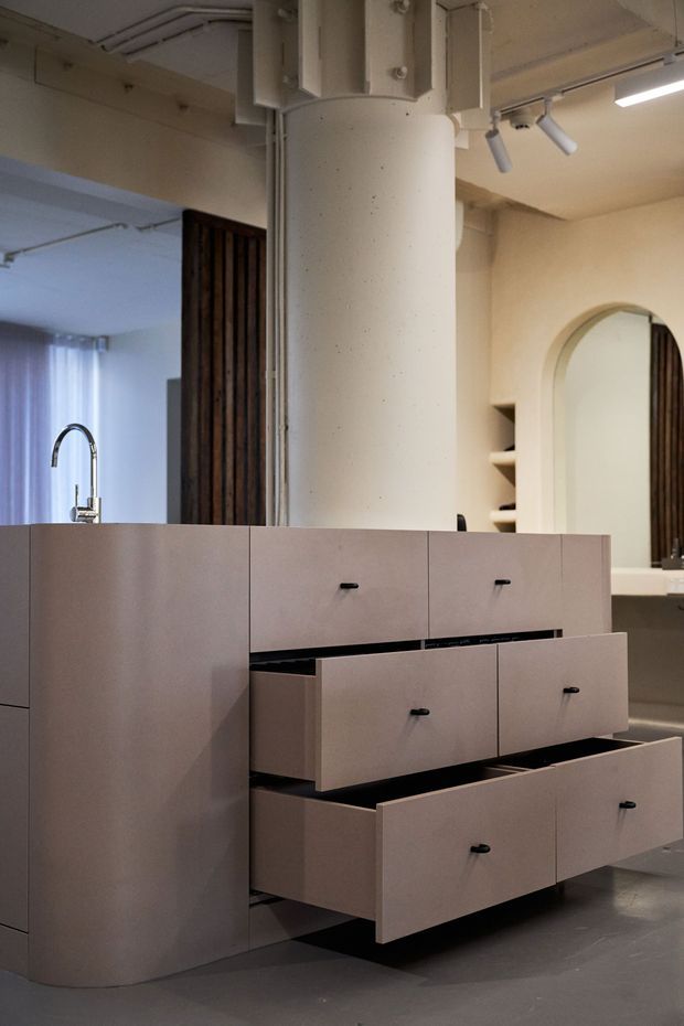 A close-up interior view of a contemporary built-in kitchen or storage island with multiple wide drawers in a muted taupe finish and small dark pulls. A stainless steel gooseneck tap is visible behind the cabinetry, while the background includes a large cylindrical structural column, exposed services, track lighting, timber slat detailing, open shelving, and an arched mirror or opening. The palette is warm neutral with dark grey flooring and a softly industrial fit-out.