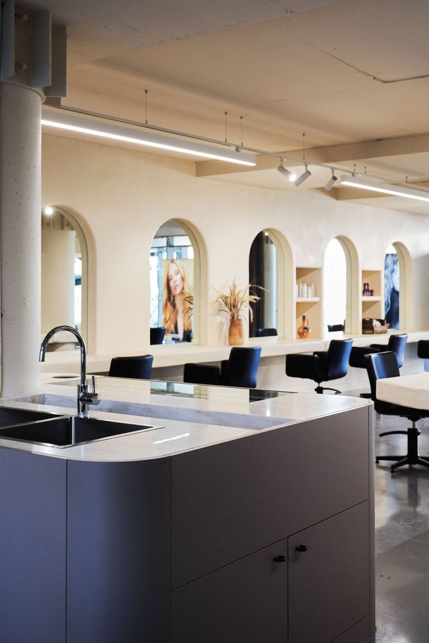 A contemporary commercial salon interior with a large dark built-in island featuring a white stone benchtop, integrated double sink and chrome gooseneck tap in the foreground. Behind it are multiple styling stations with mirrors set into repeated arched wall niches, black salon chairs, open shelving and linear suspended lighting. The space has polished concrete flooring, pale plaster walls and an industrial exposed-column and ceiling structure.
