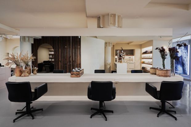 A refined salon or beauty studio reception and styling area, viewed across a long white counter with three black swivel chairs. A large wall mirror reflects the space, while dark vertical timber slats, white cabinetry and shelving, decorative dried botanicals, and warm recessed lighting create a contemporary, calm interior.