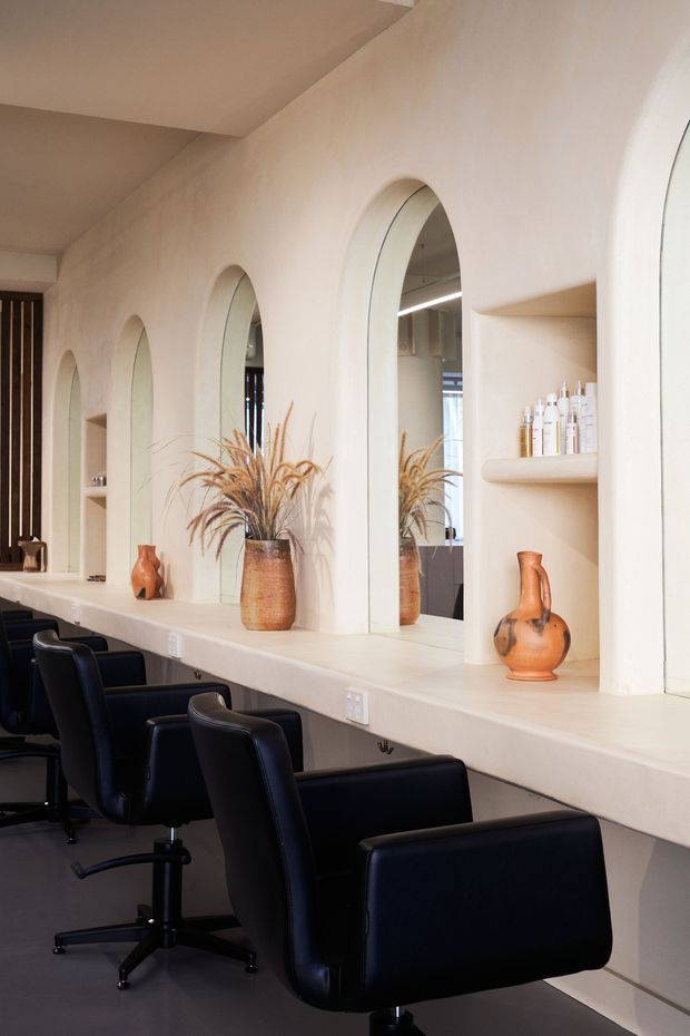 A refined salon interior with a long built-in styling counter, multiple arched mirrors, integrated wall shelves, and black leather salon chairs. The warm off-white plastered walls and countertop are accented by terracotta ceramic vases and dried grasses, creating a calm, contemporary beauty studio atmosphere.