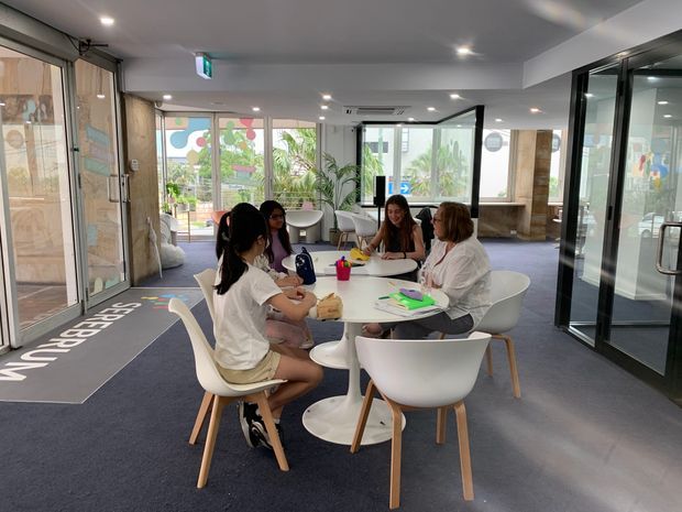 Bright contemporary communal interior with several people gathered around white round tables and molded chairs. The space has full-height glazed doors and windows, glass-partitioned rooms, recessed ceiling lighting, dark carpeted and smooth flooring, and abundant indoor planting. A visible entry mat and exit signage suggest a shared workplace, school, or commercial reception and breakout area.
