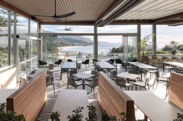 Bright contemporary café or restaurant dining area enclosed by extensive floor-to-ceiling glazing, with numerous tables, black wire-frame chairs, timber slatted booth seating, ceiling fans, and a ribbed linear ceiling. The space opens visually to a coastal bay and mountainous shoreline beyond.