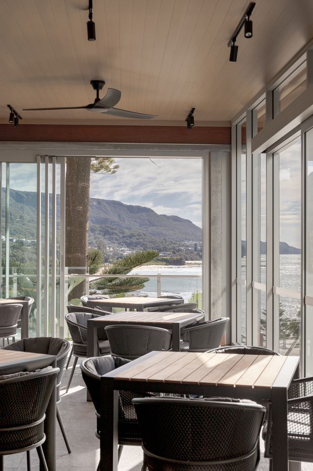 A covered waterfront dining terrace or café-style outdoor seating area enclosed with extensive glass sliding doors and windows. Multiple timber-topped tables with dark woven chairs fill the space. The ceiling has light timber lining, exposed dark structural beams, black track lighting, and a ceiling fan. A dramatic mountain, bay, and shoreline landscape is visible through the glazing.