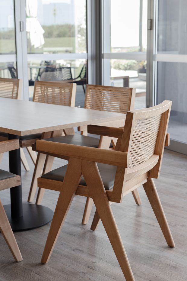 Bright contemporary dining area with a pale rectangular dining table and timber dining chairs featuring woven cane backs, black pedestal-style table legs, and a light timber-look floor. Large glazed doors or windows open the room toward an outdoor terrace, creating an airy indoor-outdoor connection.