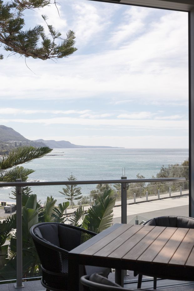 A bright coastal outdoor dining terrace overlooking the ocean and a curving shoreline. The space is framed by dark window or door edges and a glass balustrade with metal posts, with a timber slatted dining table and dark woven chairs in the foreground. Tropical and coastal planting surrounds the terrace, while a paved seaside road or promenade and distant hills are visible beyond.