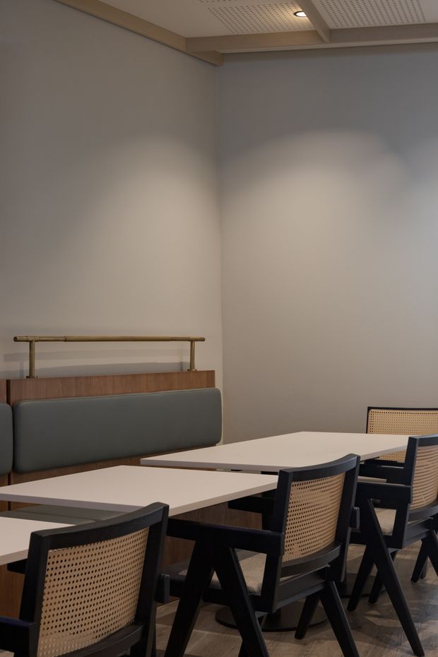 A refined interior dining or meeting area with a long light-colored table surrounded by black timber-framed chairs featuring woven cane backs. A built-in upholstered bench sits against a plain warm-grey wall, with a timber ledge and rail behind it. The ceiling has recessed lighting and perforated acoustic panels, creating a calm, contemporary hospitality or commercial setting.