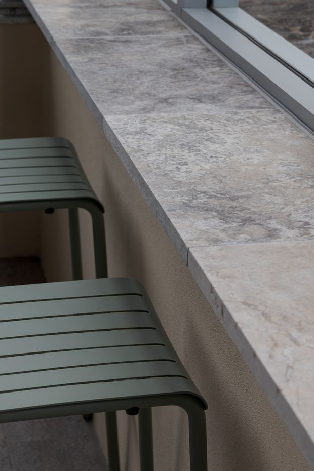 Close-up detail of a minimalist counter or bar ledge beside a large framed window, finished in a pale grey-beige marbled stone. Two dark sage-grey slatted stools sit beneath the overhang. The composition highlights the stone edge, textured wall finish, and contemporary window framing.