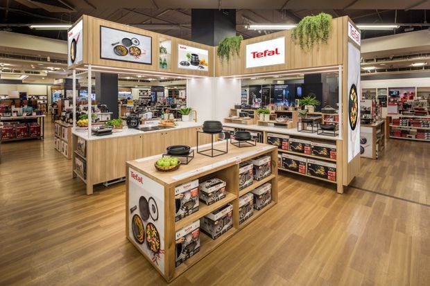 A branded Tefal retail display within a large, open commercial store. The fit-out features warm timber-look cabinetry and shelving, product displays, illuminated signage, integrated greenery, and a wood-look floor beneath an exposed dark ceiling with linear lighting.