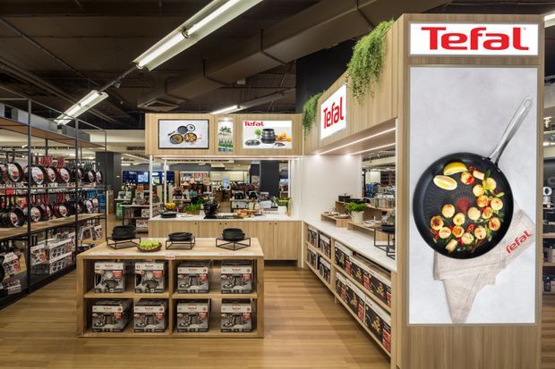 A branded Tefal retail showroom with product shelving, central display tables, illuminated signage, and promotional graphics for cookware. The space features timber-look flooring and wood-toned display joinery beneath an exposed dark ceiling with linear fluorescent lighting.