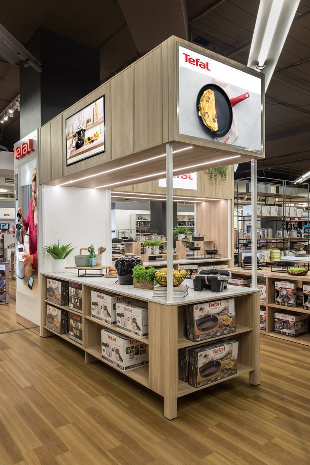 A branded Tefal retail display island inside a large store. The merchandising fixture uses light wood-look panels, illuminated shelves, product packaging, countertop cooking demonstrations, overhead advertising panels, and integrated greenery. The surrounding commercial space has exposed services, track lighting, and additional retail shelving.
