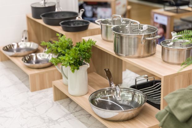 A styled kitchenware display featuring stainless-steel pots, pans, bowls, and cooking utensils arranged on tiered light-wood shelving. A white ceramic pitcher with green foliage adds a natural accent, while the pale marble-look floor and bright retail setting create a clean, contemporary presentation.