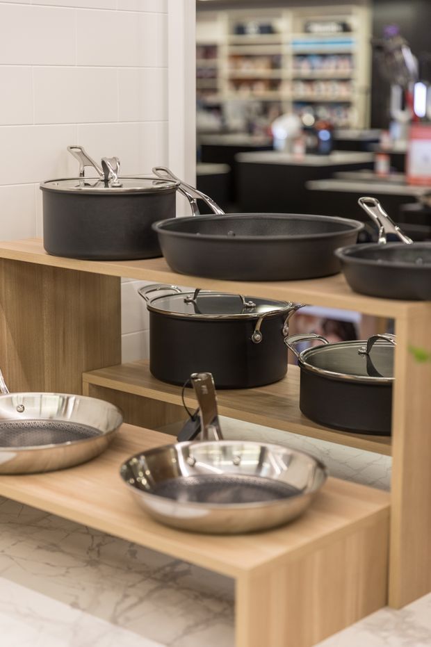 Close-up view of a cookware showroom display, featuring black and stainless-steel pots and pans arranged on light timber shelving and a counter. White tiled wall surfaces and marble-look flooring are visible, with blurred retail shelving and cabinetry in the background.