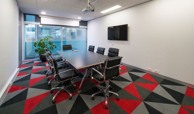 A modern commercial meeting room with a long dark conference table surrounded by black leather swivel chairs. The room has large glazed windows, a wall-mounted display, ceiling-mounted projector, recessed linear lighting, and a bold geometric carpet in red, charcoal, grey, and black. The space is functional, contemporary, and brightly lit.