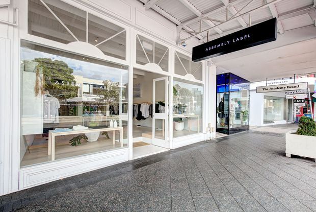 Bright covered retail arcade with a white-painted shopfront for Assembly Label. The storefront features large glazed display windows, white framed doors and transom windows, decorative geometric glazing bars, and a white exposed structural ceiling. Dark tiled paving runs through the pedestrian arcade, with adjacent retail shops visible in the background.