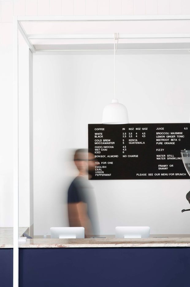 Bright minimalist café interior viewed through a large glazed service opening. A dark navy counter fronts the space, with a pale stone or terrazzo benchtop, white pendant lighting, white chairs, and a black menu board listing coffee, juice, and brunch items. A blurred staff member stands behind the counter. White vertical wall panelling and exposed white framing create a clean, modern commercial fit-out.