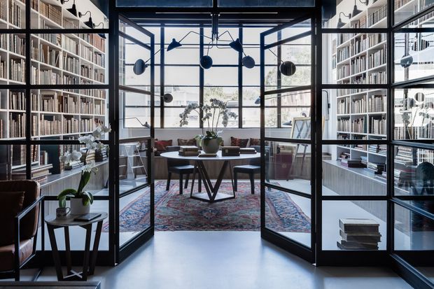 A refined home library and study viewed through black steel-framed glass doors. Floor-to-ceiling bookshelves line both sides, while a central desk with chairs sits on a patterned rug in front of expansive industrial-style glazing. A sculptural pendant light, indoor plants, and curated books complete the sophisticated, light-filled space.