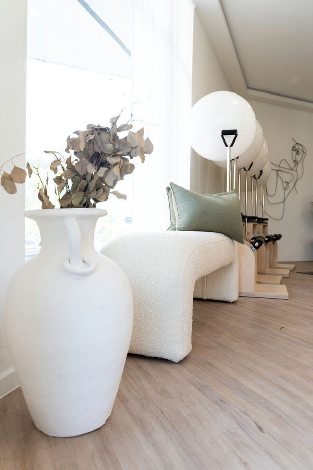 A bright, refined living room with pale timber flooring, a sculptural cream boucle armchair, a muted sage cushion, and a large white ceramic vase holding dried foliage. A globe floor lamp, minimalist sculptural wall art, and open shelving add understated contemporary character. The room has abundant natural light from large sheer-covered glazing.