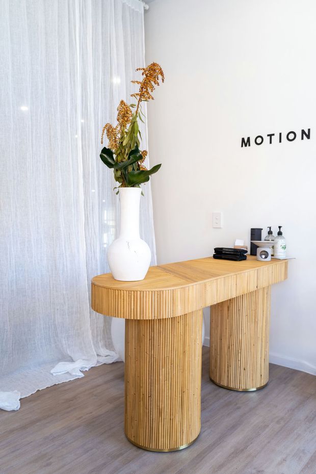 Bright minimalist interior vignette featuring a curved fluted timber console table with rounded cylindrical supports, placed against a white wall beside sheer floor-to-ceiling curtains. A white ceramic vase with dried foliage, small black accessories, and the word “MOTION” displayed on the wall add restrained decorative detail. The floor is a muted wood-look finish.