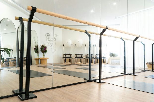 Bright, spacious yoga or dance studio with full-height mirrored walls, a pale timber floor, and freestanding black metal ballet barre frames with timber rails. The mirrored reflection reveals exercise mats, candles, plants, and a light-filled adjoining area.