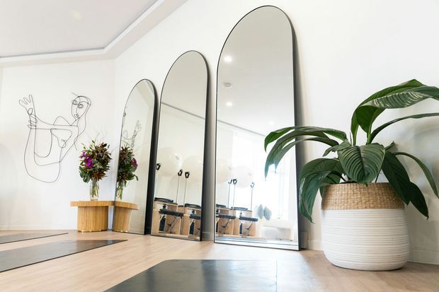 Bright, refined interior featuring three tall arched mirrors with dark frames, pale timber flooring, large potted plants, floral arrangements, and a minimalist line-art wall drawing. Reflections reveal exercise equipment, suggesting a boutique fitness or Pilates studio. The room has a high-quality, airy fit-out with a recessed ceiling detail and neutral finishes.
