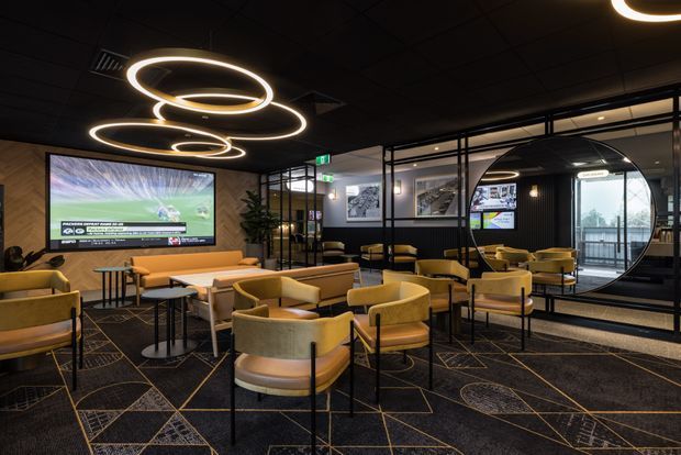 A stylish commercial lounge or hospitality space with numerous mustard and tan upholstered lounge chairs, small tables, dark patterned carpet, black ceilings and walls, extensive black-framed glass partitions, and a large wall-mounted television displaying sports. Sculptural glowing ring pendant lights form a strong focal point overhead, while a circular glazed opening and reflective surfaces add visual interest.