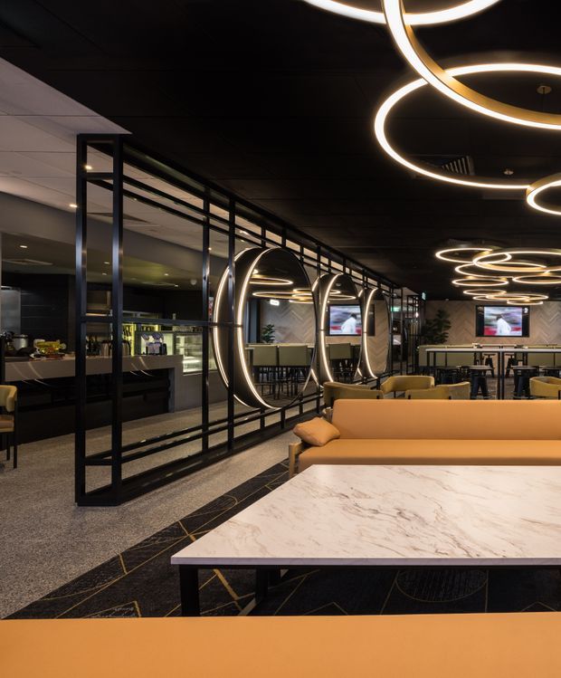 A sophisticated commercial hospitality lounge or restaurant interior with a dark ceiling, black metal-framed glass partitions, integrated bar or dining area, mustard and tan upholstered seating, and a large pale marble-effect communal table. Sculptural circular ring pendant lights create a dramatic illuminated ceiling feature, while wall-mounted screens and reflective curved details add a contemporary upscale atmosphere.