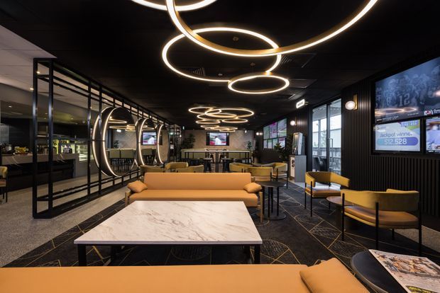 A sophisticated contemporary hospitality lounge or sports betting venue with a dark interior, black ceiling and walls, warm mustard-yellow upholstered sofas and armchairs, marble-look tables, and dramatic circular LED pendant lighting. Glazed partitions and mirrors create depth, while wall-mounted screens and betting displays indicate a commercial entertainment space.