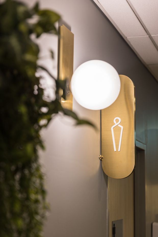 Close-up architectural detail of a restroom or bathroom entry sign mounted on a wall. The sign has a warm brushed-gold metal panel with a white female pictogram and a spherical illuminated globe light above it. A blurred leafy plant occupies the foreground, with a glass doorway or partition and suspended ceiling visible to the right and above. The image has a refined, softly lit commercial-interior character.
